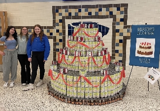 three female students next to a giant cake made of cans