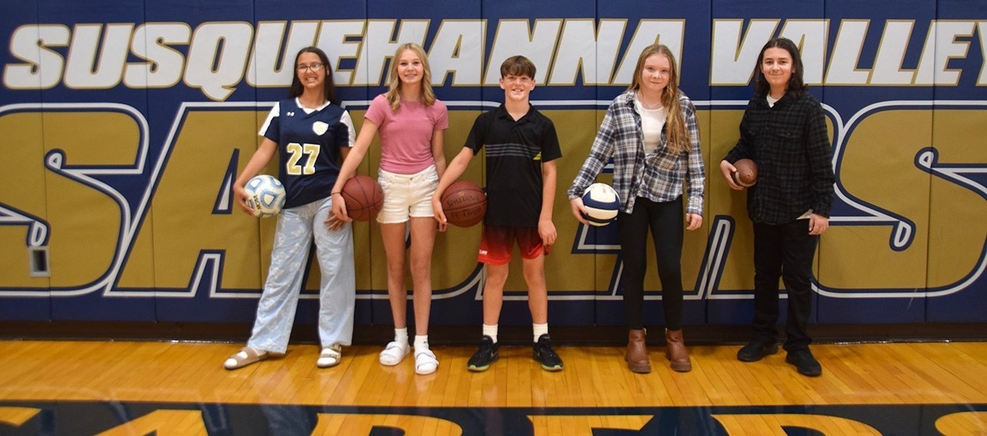 Five Richard Stank middle school students holding sports balls stand by the Susquehanna Valley Sabers wall in the gym.