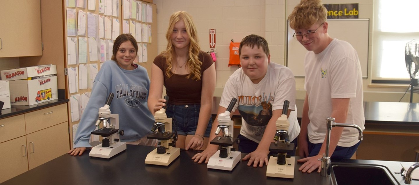 Four middle school students smile at the camera as they appear to be using microscopes in science class.